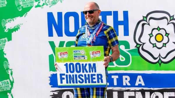 A man wearing blue and yellow Doddie tartan shorts and a My Name'5 Doddie Foundation blue and yellow tartan t-shirt is standing up, smiling and holding a sign which says '100km Finisher'. He has medals around his neck as well and is standing in front of a large sign which says 'North Yorks Ultra Challenge'.