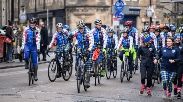 A group of cyclists dressed in blue and yellow tartan clothing cycling down into Edinburgh's Grassmarket Doddie Aid raises £1.35 million as total funds committed to MND research nears £20 million milestone