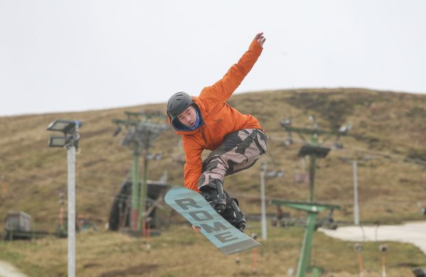 A man in an orange jacket holds a snowboard at a ski slope with scenic hills in the background.