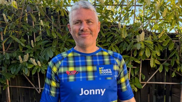 Jonathan Beaton standing outside in his garden in the sunshine, looking at the camera and smiling. He is wearing a Doddie Weir blue and yellow tartan running top.