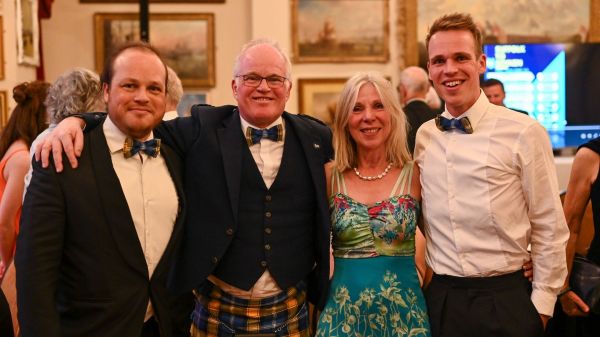 Nick Apperely and his family at a black tie ball fundraising event which Nick's son Harry organised to fundraise for My Name'5 Doddie Foundation. There are four people looking at and smiling at the camera, with their arms around each other. From left to right there is Nick's son Josh who is wearing a blue and yellow Doddie tartan bow tie, Nick who is wearing a blue and yellow Doddie tartan kilt and bowtie, Nick's wife Jill who is wearing a blue, pink and green floral dress, and Nick's son Harry, who is also wearing a blue and yellow Doddie tartan bowtie.