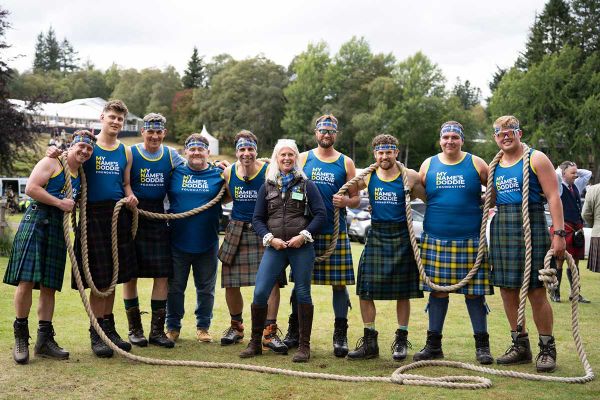 A group in kilts and matching shirts poses with ropes for a charity event outdoors.