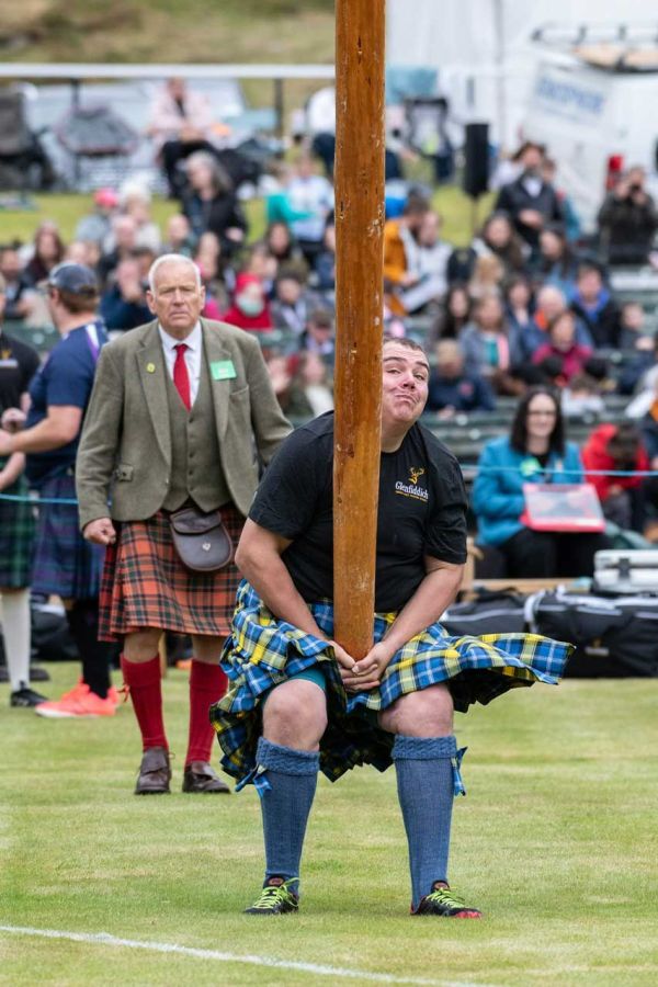 A man in a kilt prepares to lift a wooden pole at a crowd-filled event.