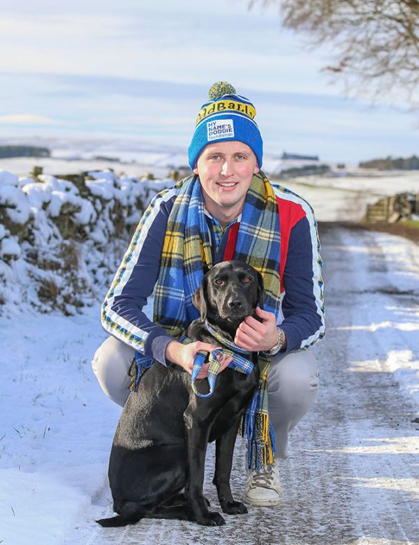 A man in winter clothes crouches beside a black dog on a snow-covered path.