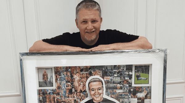 Steven Hart, an artist living with MND, is standing behind one of his portraits, of Scott Kyle, and resting his forearms on the top of the portrait's frame. Steven is smiling at the camera.